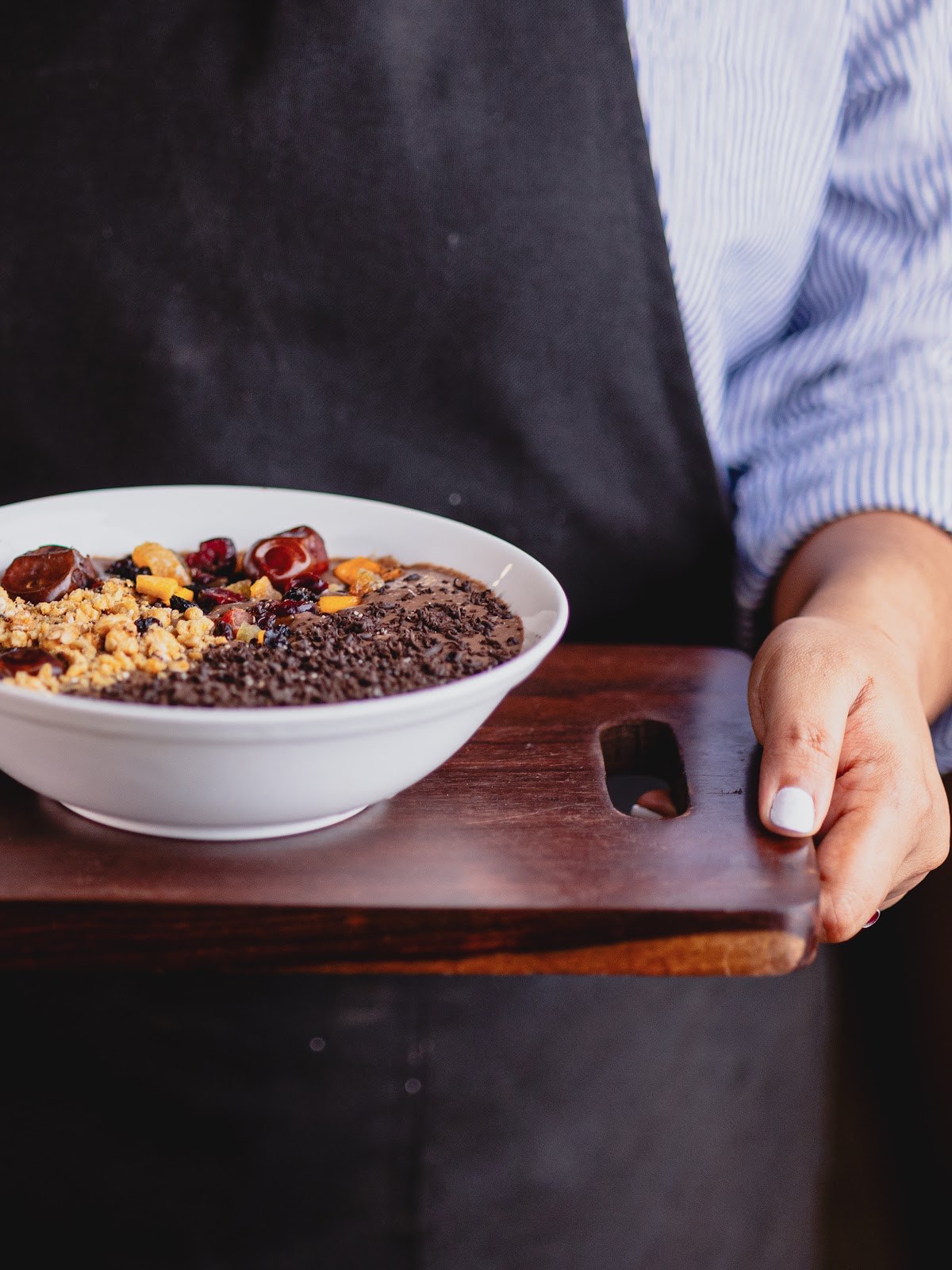 A server carrying a bowl of ragi porridge topped with dates and granola on a dark wooden board
