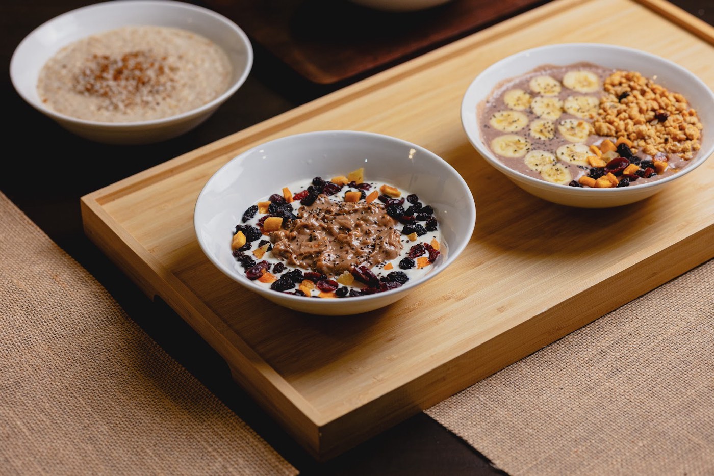 Three breakfast bowls on a wooden board — oatmeal, a yogurt bowl with peanut butter and dried fruit, and a banana smoothie bowl with granola