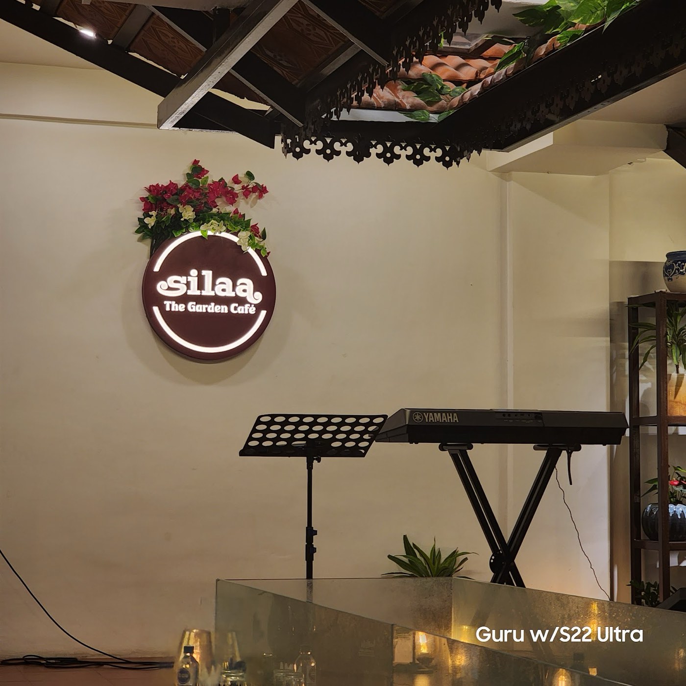 The illuminated Silaa roundel on a cream wall with bougainvillea above it, a Yamaha keyboard and music stand set up beneath