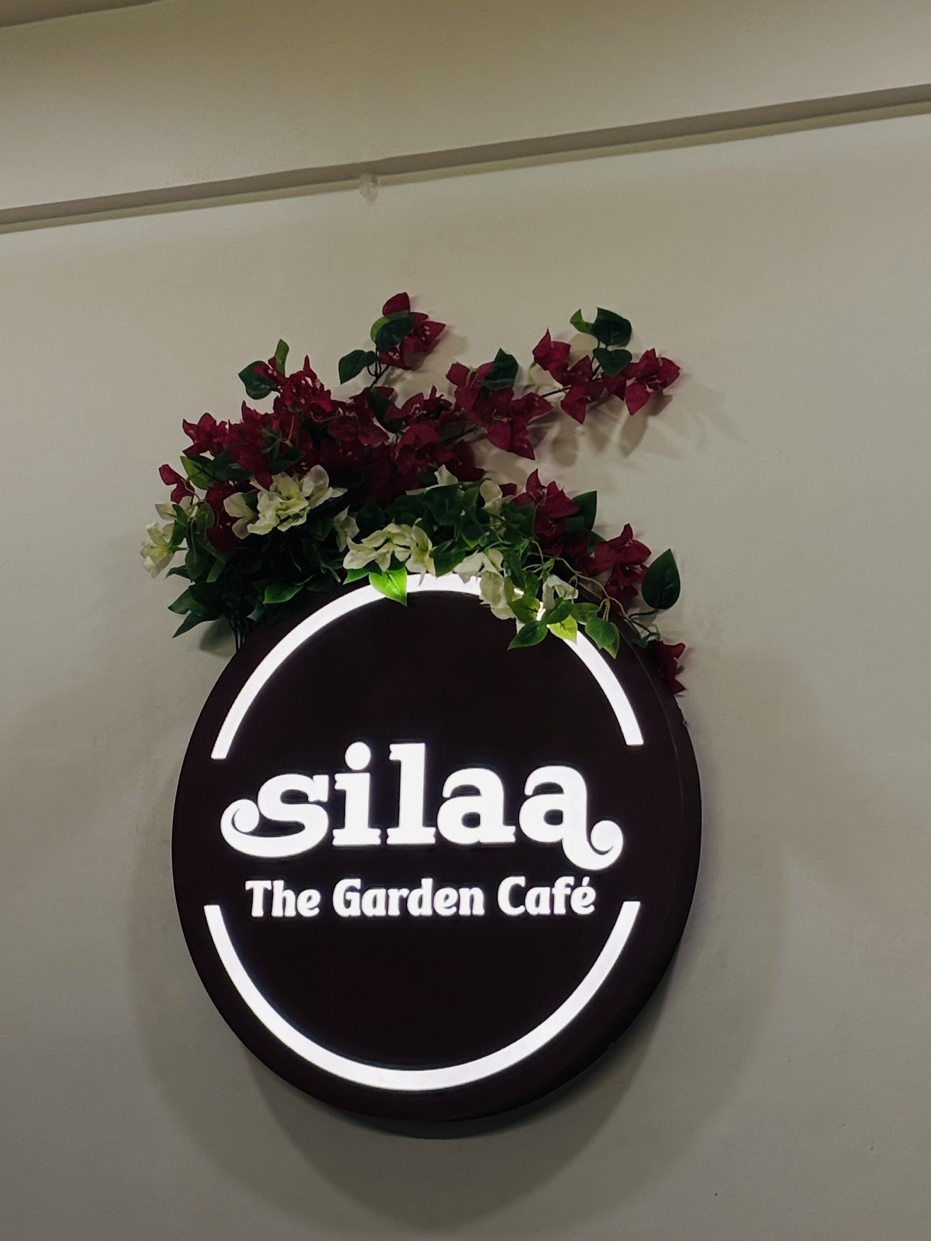The Silaa — The Garden Café roundel sign, dark with white lettering, crowned with bougainvillea